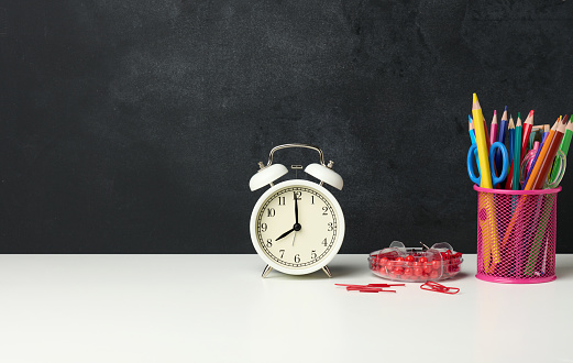 white round alarm clock and a metal glass with pens, pencils and felt-tip pens on the background of an empty black chalk board, copy space