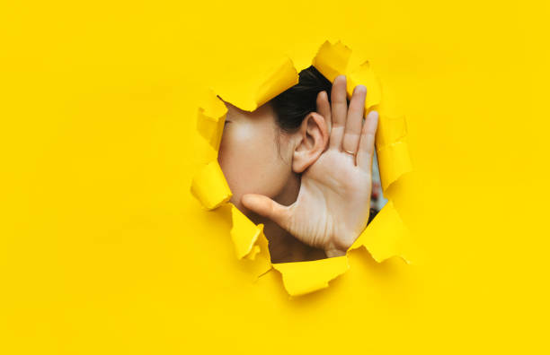 close-up of a woman's ear and hand through a torn hole in the paper. yellow background, copy space. the concept of eavesdropping, espionage, gossip and tabloids. - communication photography part of torn imagens e fotografias de stock