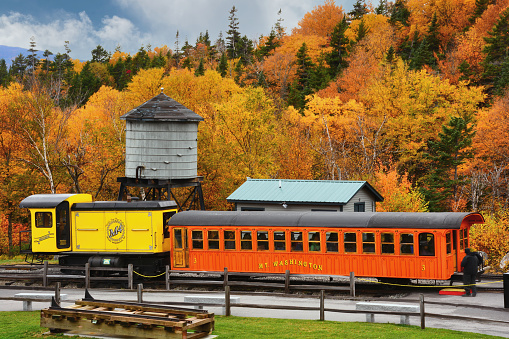 Base Station at Mount Washington Cog Railway – October 19, 2016: In operation since 1886, the Mount Washington Cog Railroad is a popular destination for tourists visiting New Hampshire. The modern biodiesel locomotive and vintage passenger car parked here at the loading dock transport passengers to the summit of Mount Washington. The nearby water tank is used for older coal-fired steam locomotives still in use by the railway.