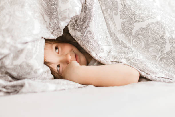 4 세의 귀여운 소년이 담요 아래에서 밖으로 보입니다. 집에서 아침 재미. 삶의 순간을 선물합니다. 아이들과 함께 침대에서 놀기 - portrait germany party bed 뉴스 사진 이미지