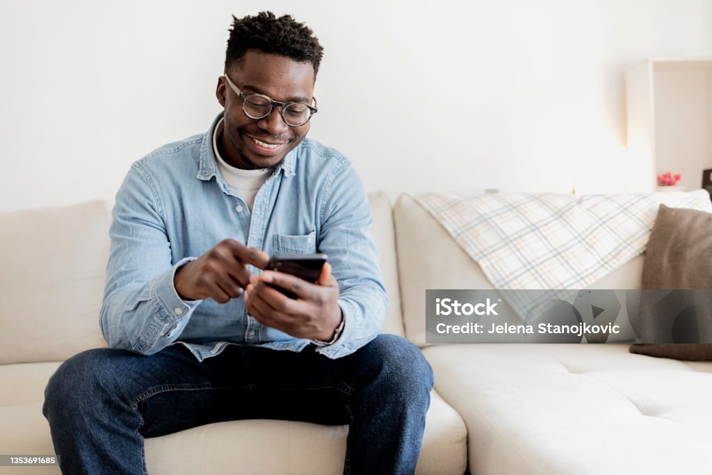 Cheerful black man using mobile wearing eyeglasses, texting and browsing internet. Portrait of an African American man at home texting on his cellphone while sitting on the sofa - lifestyle concept. Cheerful black man using mobile wearing eyeglasses, texting and browsing internet. Using Phone Stock Photo