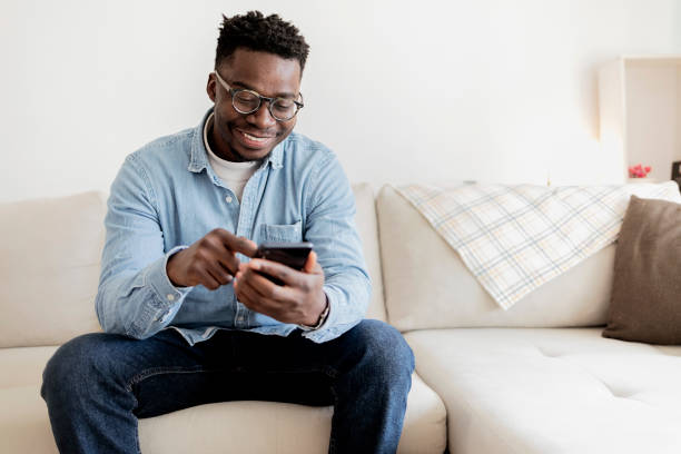 Cheerful black man using mobile wearing eyeglasses, texting and browsing internet. Portrait of an African American man at home texting on his cellphone while sitting on the sofa - lifestyle concept. Cheerful black man using mobile wearing eyeglasses, texting and browsing internet. guy looking at cell phone stock pictures, royalty-free photos & images