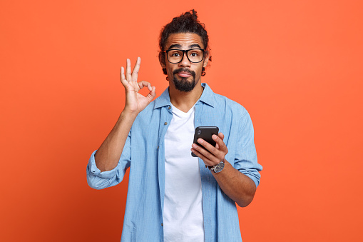 Isolated photo of young african american man in eyeglasses showing ok sign with mobile phone in hand and looking at camera, guy demonstrating gesture of approval while reading good news on smartphone