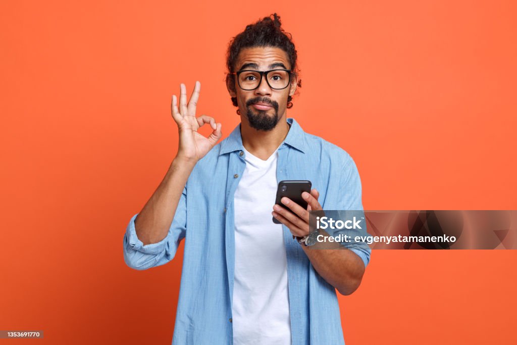 Isolated studio shot of young african american man showing ok sign with mobile phone in hand Isolated photo of young african american man in eyeglasses showing ok sign with mobile phone in hand and looking at camera, guy demonstrating gesture of approval while reading good news on smartphone Men Stock Photo