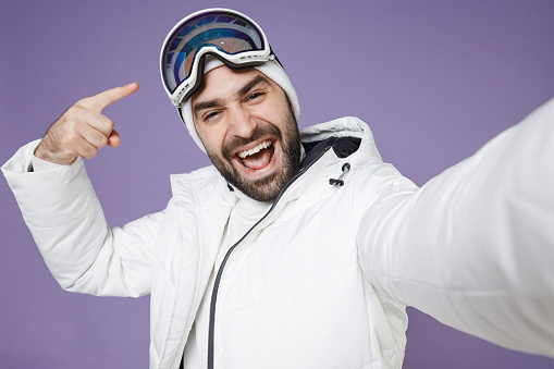Close up of funny skier man in windbreaker jacket pointing finger on ski goggles mask doing selfie shot on mobile phone spend weekend in mountains isolated on purple background. People hobby concept