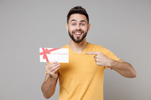Young caucasian smiling bearded attractive handsome happy man 20s wearing casual yellow basic t-shirt point index finger on gift voucher flyer mock up isolated on grey color background studio portrait