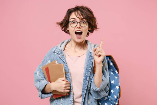 Excited girl student in denim clothes glasses backpack isolated on pastel pink background. Education in high school university college concept. Hold books pointing index finger up with great new idea. Excited girl student in denim clothes glasses backpack isolated on pastel pink background. Education in high school university college concept. Hold books pointing index finger up with great new idea isolated color stock pictures, royalty-free photos & images