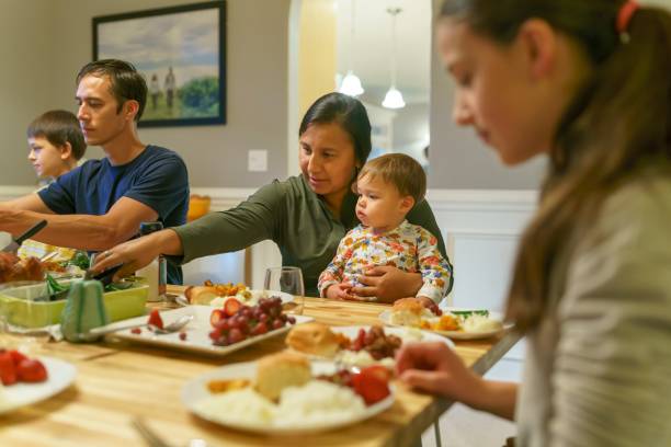 Family eating together at holiday table Multi-ethnic group of adults and children of Eurasian, Native American and Caucasian descent sitting around a dining room table filled with food for a traditional winter holiday dinner serving themselves and eating while celebrating with family and friends. Selective focus on a Native American woman serving herself green beans while holding a mixed race toddler girl in her lap. indian dinner stock pictures, royalty-free photos & images