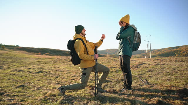 Charming boyfriend, proposing his girlfriend with a ring, during their autumn hike in the sunset, while kneeling on one knee