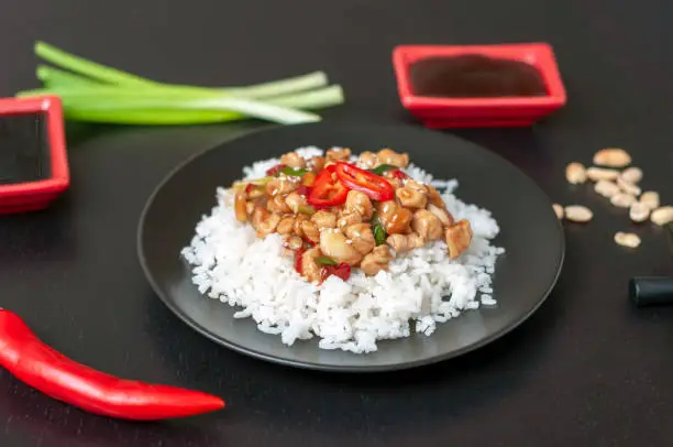 Photo of Chicken Kung pao or gongbao on rice plates on a black table with traditional sauces and ingredients. A popular dish of Sichuan cuisine of China made of chicken, peanuts, hot pepper, sauces and spices