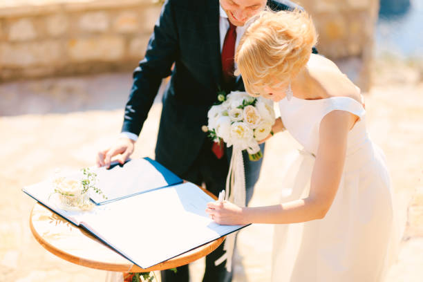 le marié en costume noir et la mariée en robe blanche avec un bouquet de fleurs signent le livre d’enregistrement du mariage - traditional ceremony sign symbol wedding photos et images de collection