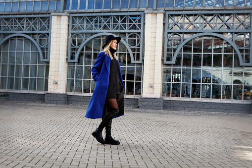 Fashionable beautiful woman walking on the street. Stylish trend in autumn or spring clothes is blue coat, felt hat, black leather FLAT KNEE HIGH BOOTS and black shorts