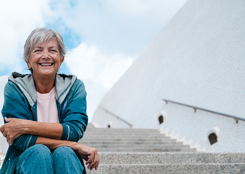 Portrait of attractive senior woman sitting outdoors on staircases looking at camera smiling. Cloudy sky on background