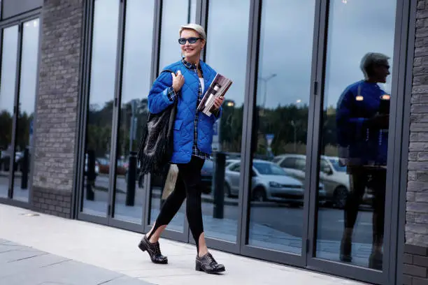 Fashionable woman with short hair walking street dressed in stylish casual outfit, blue colorful jacket and sunglasses, black leather handbag and boots, leggings with straps. Trend spring or autumn