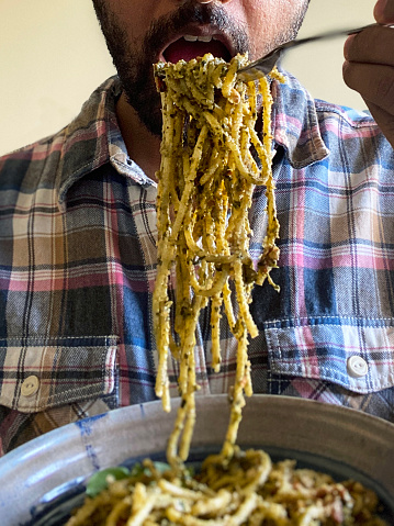 Stock photo showing an Indian man about to eat a forkful of homemade spaghetti pesto sauce pasta from a blue and white dish.