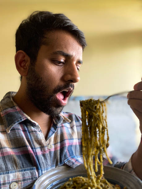 close-up image of indian man holding metal fork of pesto, spaghetti, eating pasta topped with homemade pesto sauce, mouth open, focus on foreground - spaghetti cooked heap studio shot imagens e fotografias de stock