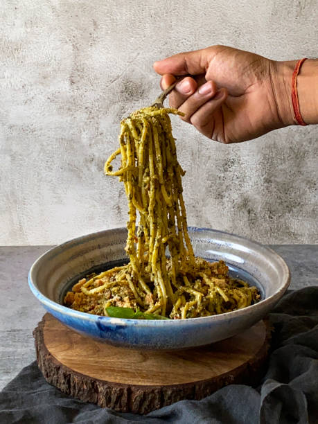 image of metal fork twirling pesto, spaghetti held in hand of unrecognisable person, pasta topped with homemade pesto sauce, portion on blue and white dish garnished with basil leaves, elevated view, focus on foreground - spaghetti cooked heap studio shot imagens e fotografias de stock