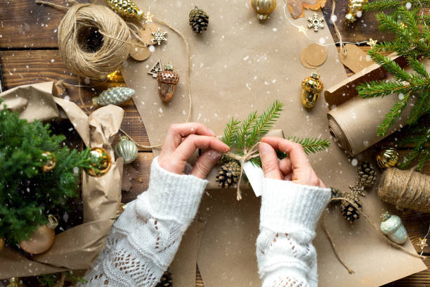 istock Hands in a white sweater pack a gift for Christmas and new year in eco-friendly materials: kraft paper, live fir branches, cones, twine. Tags with mock up, natural decor, hand made, DIY. Festive mood 1353760156