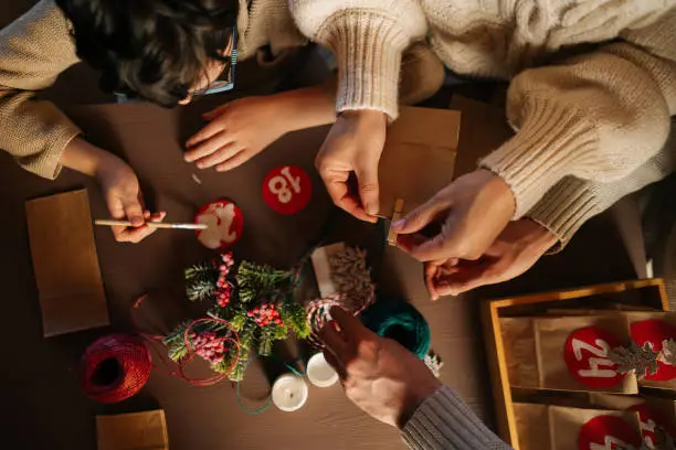 High angle view of unrecognizable family making Christmas advent calendar together sitting at table in house on xmas Eve. Father, mother and son enjoying togetherness preparing for holidays at home.