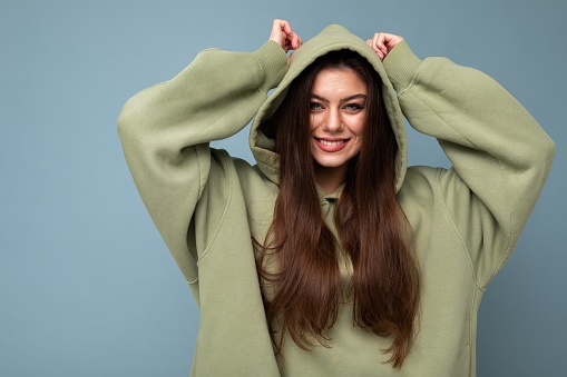 Portrait of attractive young caucasian smiling brunette woman model in trendy khaki hoodie and wearing hood isolated on blue background with copy space. Positive concept.