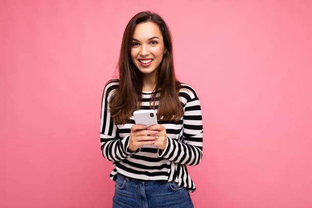 istock smiling Beautiful young brunette woman using mobile phone communicating via texting message wearing sweater isolated on wall background looking at camera 1353775874