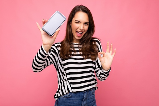 Winking beautiful happy young woman wearing striped sweater isolated over background with copy space showing ok gesture looking at camera showing mobile phone screen display. Mock up, cutout