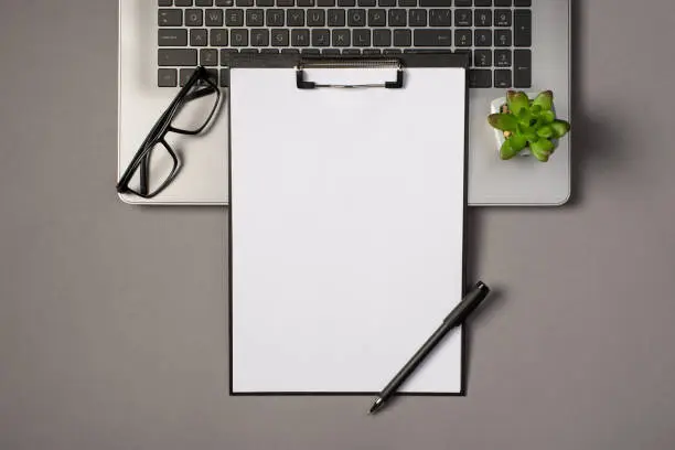 Photo of Top view photo of black clipboard folder with white paper sheet pen glasses and plant on grey laptop keyboard on isolated grey background with copyspace