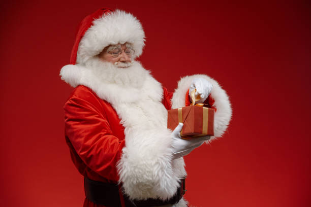 istock Santa Claus is holding a little gift box, standing on a red background 1353786053