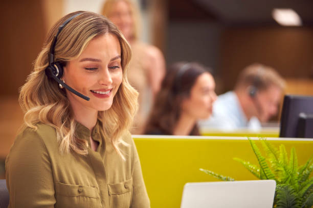 Businesswoman In Cubicle Wearing Headset Talking To Caller In Busy Customer Services Centre stock photo
