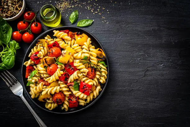 Vegan food and healthy eating: overhead view of a pasta salad plate shot on black table. Cherry tomatoes, basil leaves and an olive oil bottle complete the composition. The composition is at the left of an horizontal frame leaving useful copy space for text and/or logo at the right. High resolution 42Mp studio digital capture taken with SONY A7rII and Zeiss Batis 40mm F2.0 CF lens
