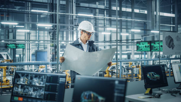 istock Engineer in Uniform and Hard Hat Looking at a Technical Blueprint at Work in an Office at Car Assembly Plant. Industrial Specialist Working on Vehicle Parts in Technological Development Facility. 1353796788