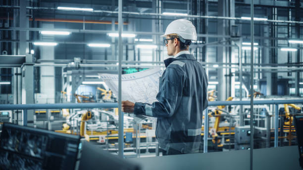 istock Engineer in Uniform and Hard Hat Looking at a Technical Blueprint at Work in an Office at Car Assembly Plant. Industrial Specialist Working on Vehicle Parts in Technological Development Facility. 1353796805