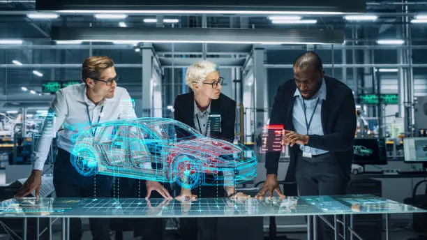 Photo of Diverse Team of Engineers Working in Office at Industrial Factory. Industrial Designers Discuss Electric Car Augmented Reality Hologramm. Specialists Work in Technological Car Development Facility.