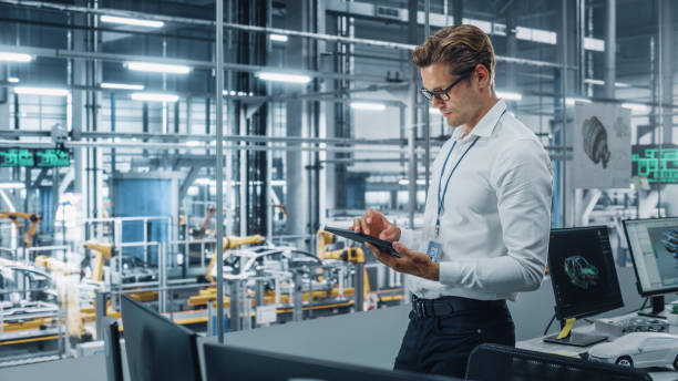 istock Handsome Engineer in Glasses and White Shirt Using Tablet Computer and Looking Out of the Office at a Car Assembly Plant. Industrial Specialist Working on Vehicle Design in Technological Facility. 1353796918