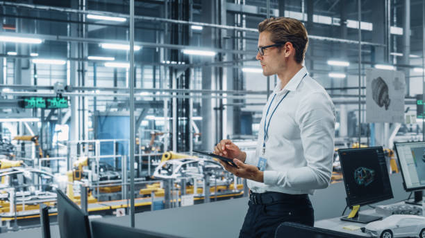 istock Handsome Engineer in Glasses and White Shirt Using Tablet Computer and Looking Out of the Office at a Car Assembly Plant. Industrial Specialist Working on Vehicle Design in Technological Facility. 1353796950