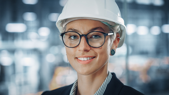 Portrait of a Happy Smiling Young Beautiful Female Engineer Wearing Glasses and White Hard Hat in Office at Car Assembly Plant. Industrial Specialist Working on Vehicle Design in Modern Facility.