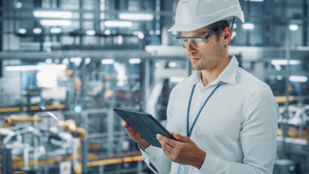 istock Male Engineer in Safety Goggles and Hard Hat Using Tablet Computer and Looking Out of the Office at a Car Assembly Plant. Industrial Specialist Working on Vehicle Production in Technological Factory. 1353796972