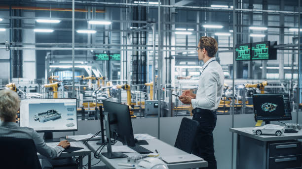 istock Handsome Engineer in Glasses and White Shirt Using Tablet Computer and Looking Out of the Office at a Car Assembly Plant. Industrial Specialist Working on Vehicle Design in Technological Facility. 1353796983