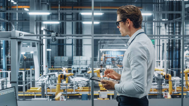 istock Handsome Engineer in Glasses and White Shirt Using Tablet Computer and Looking Out of the Office at a Car Assembly Plant. Industrial Specialist Working on Vehicle Design in Technological Facility. 1353797016