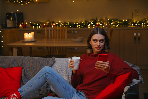 A  young cute woman with bob haircut in a red sweater sits on a cozy gray sofa with bright red cushions and drinks tea,  holding her red smartphone, looking away with a mysterious smile, we see her half-seated with Christmas lights and candles behind her