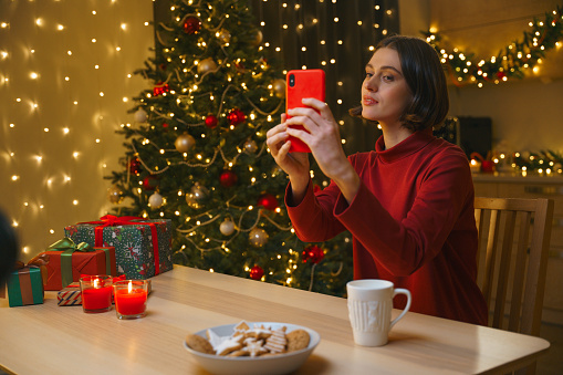 A young pretty female blogger makes selfie on her red smart phone in a Christmas interior, we see her on a kitchen with the Christmas tree and table with cookies and a cup of herbal tea