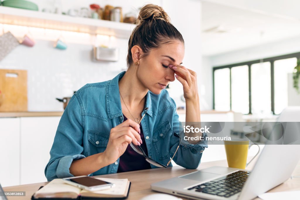 Stressed business woman working from home on laptop looking worried, tired and overwhelmed int he kitchen at home. Shot of stressed business woman working from home on laptop looking worried, tired and overwhelmed int he kitchen at home. Women Stock Photo