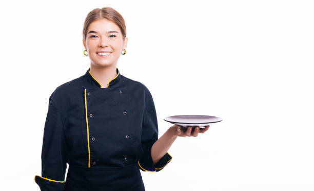 istock Joyful young woman chef standing over white background and holding empty black plate 1353821536