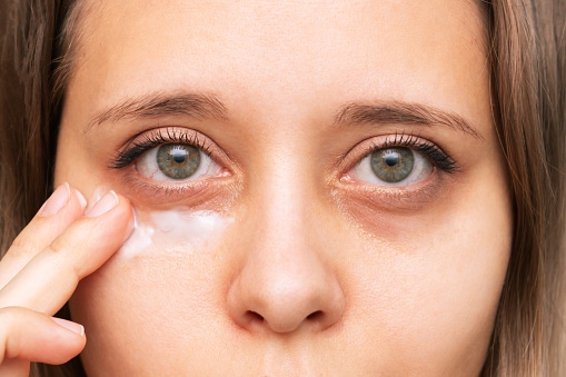 Una mujer joven tocándose una piel de la cara mientras se aplica la crema para los ojos negros photo