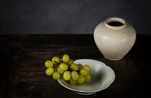 Still life of Chinese antique porcelains, a white jar and a white flower shaped plate, with branch of white grapes on wooden table against gray background