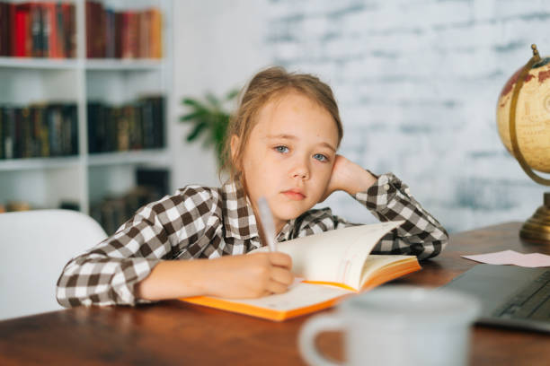 istock Front view of bored tired cute elementary child school girl doing writing homework holding pen sitting at table with laptop and paper notebook at home 1353835154