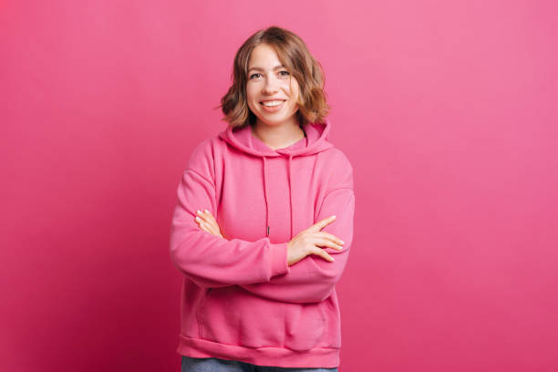 Photo of young smiling woman with crossed arms in pink hoodie standing over pink background stock photo