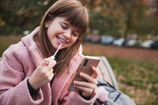 Happy young girl wearing dental braces applying lip-gloss when sitting on a park bench and using phone as a mirror