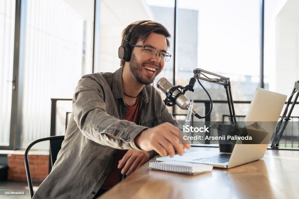 Laughing man host streaming podcast in studio Young man host in headphones and glasses enjoying podcasting in studio, speaking into a microphone, holding a pen, using laptop. Handsome podcaster laughing while streaming live audio podcast Podcasting Stock Photo