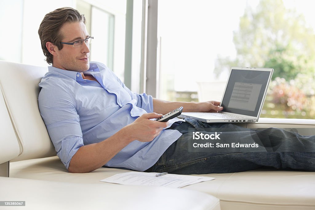 Uomo disteso sul divano con il telecomando e laptop - Foto stock royalty-free di Guardare la TV